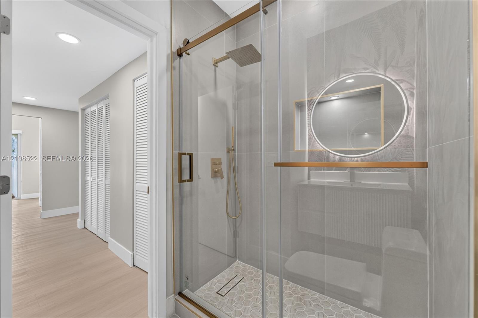 Walk-in shower featuring a frameless glass enclosure, gold-tone hardware, a rainfall showerhead, and a tiled hexagonal floor pattern, paired with a modern floating vanity and a circular back-lit mirror