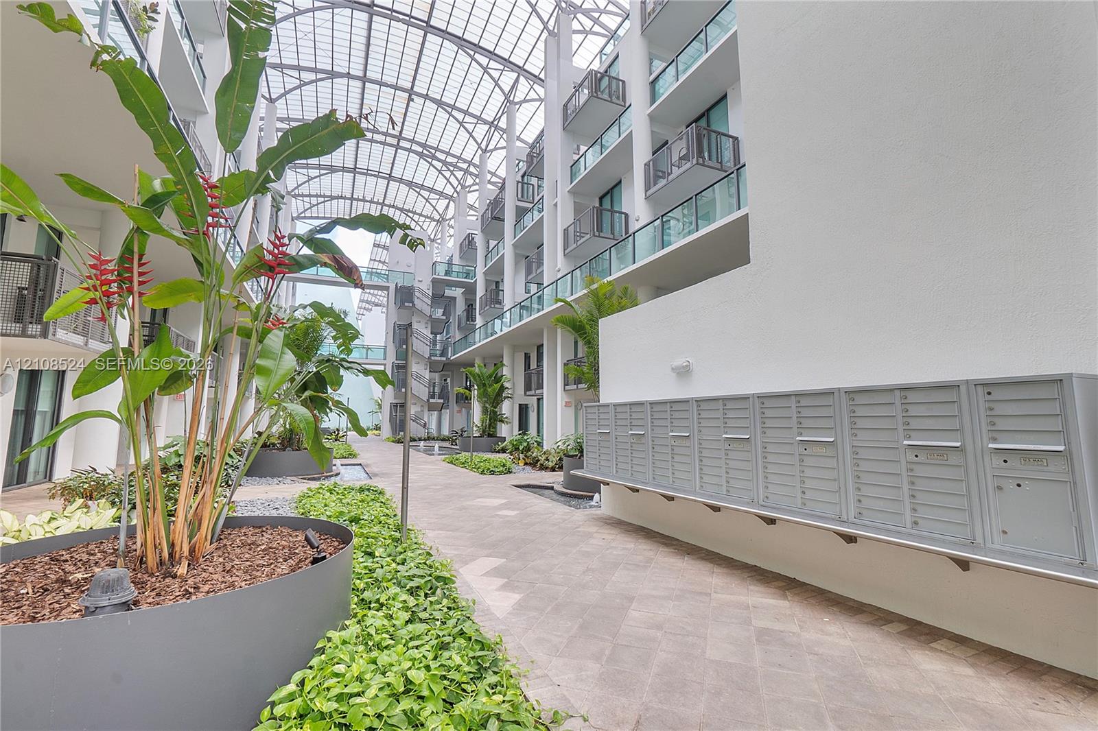 Atrium courtyard featuring a vaulted glass-paneled ceiling, herringbone-patterned stone pavers, metal cluster mailbox unit, and integrated landscape planters