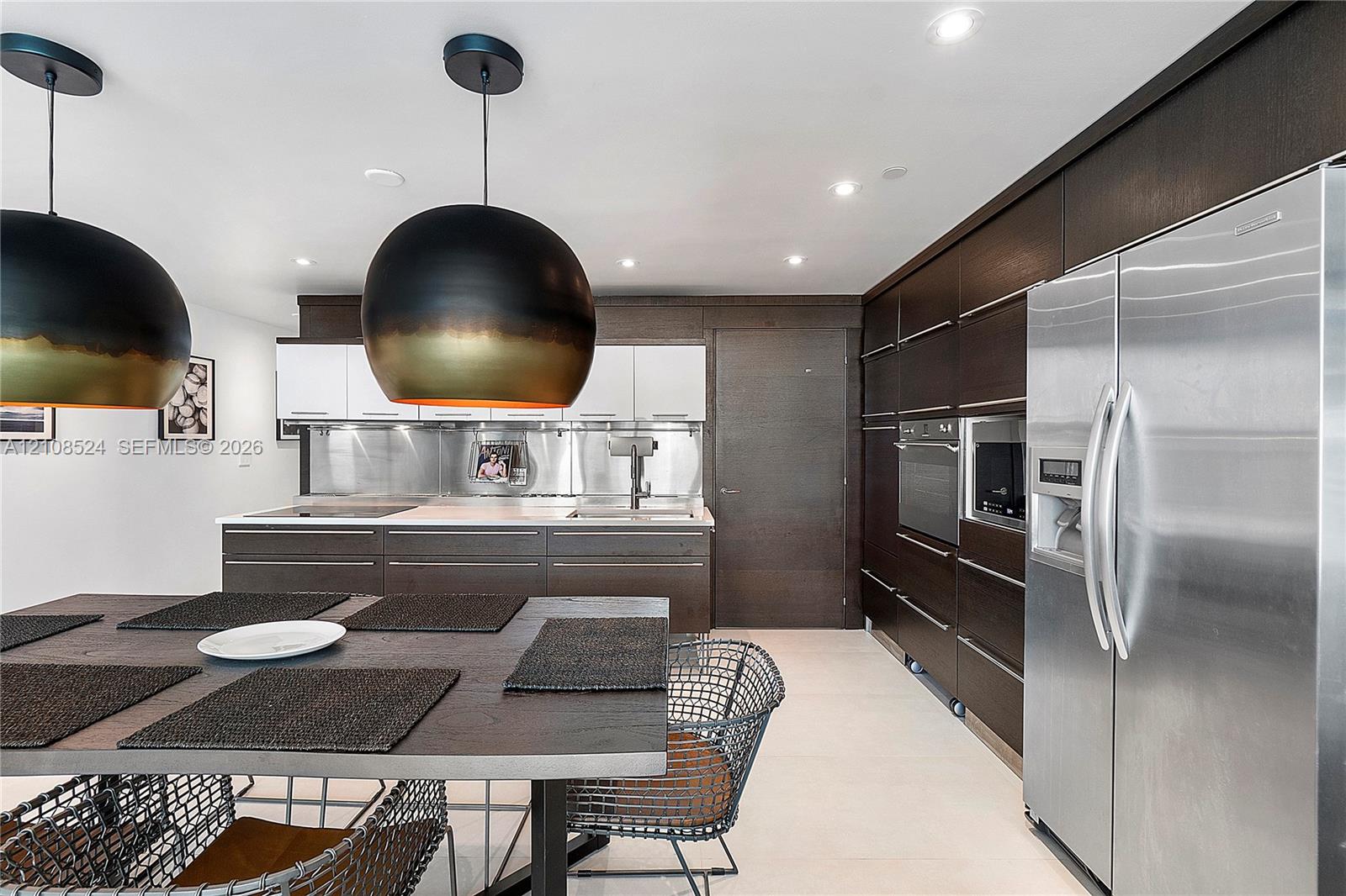Modern kitchen featuring dark wood-finish cabinetry, a stainless steel refrigerator with a built-in wall oven and microwave, metal backsplash, and oversized dome pendant lighting