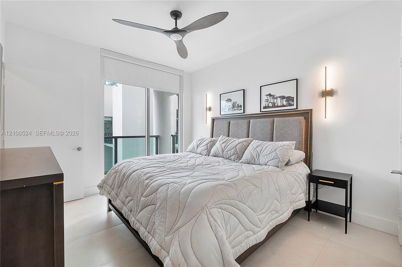 Minimalist interior featuring large-format tile flooring, dual vertical wall-mounted sconces, a matte black three-blade ceiling fan, and floor-to-ceiling glass sliding doors with access to a railed balcony Minimalist interior featuring large-format tile flooring, dual vertical wall-mounted sconces, a matte black three-blade ceiling fan, and floor-to-ceiling glass sliding doors with access to a railed balcony
