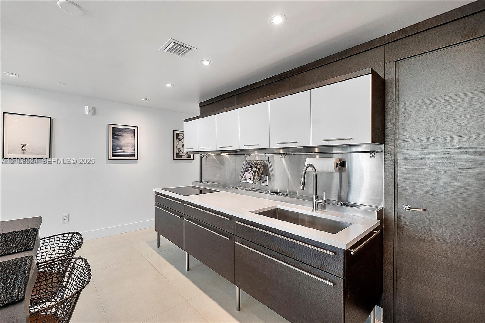 Kitchen featuring white and dark wood-finish cabinetry, light stone-surface countertops, stainless steel backsplash, integrated cooktop, and undermount stainless steel sink