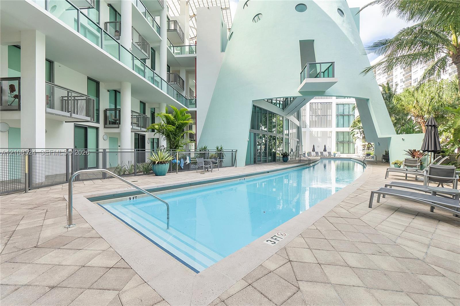 In-ground swimming pool with stepped entry, expansive stone-paver decking, and prominent architectural sculptural facade featuring metal railings and glass balcony enclosures