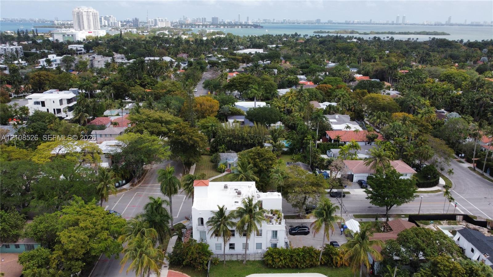 Aerial perspective of a multi-story, white stucco structure featuring a flat roofline, integrated balconies, and established perimeter landscaping situated within a tree-lined residential setting