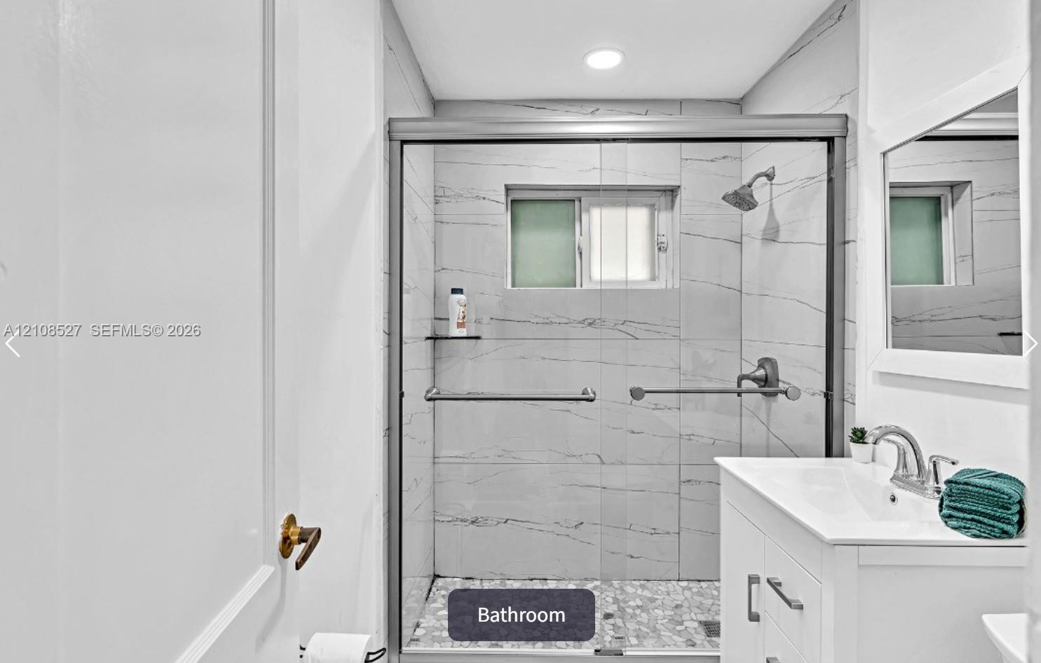 Bathroom featuring a walk-in shower with stone-patterned wall tile, a glass sliding enclosure, pebble-tile flooring, and a white vanity with a polished metal faucet