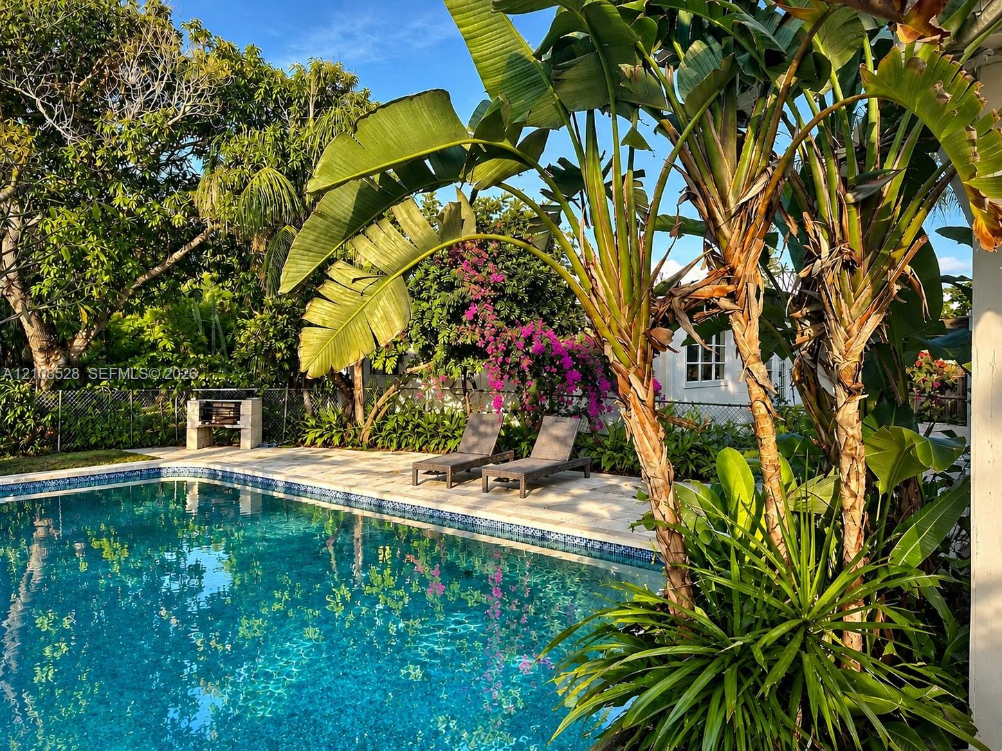 In-ground swimming pool with tiled water-line, stone paver patio, and built-in masonry barbecue station, complemented by mature tropical landscaping