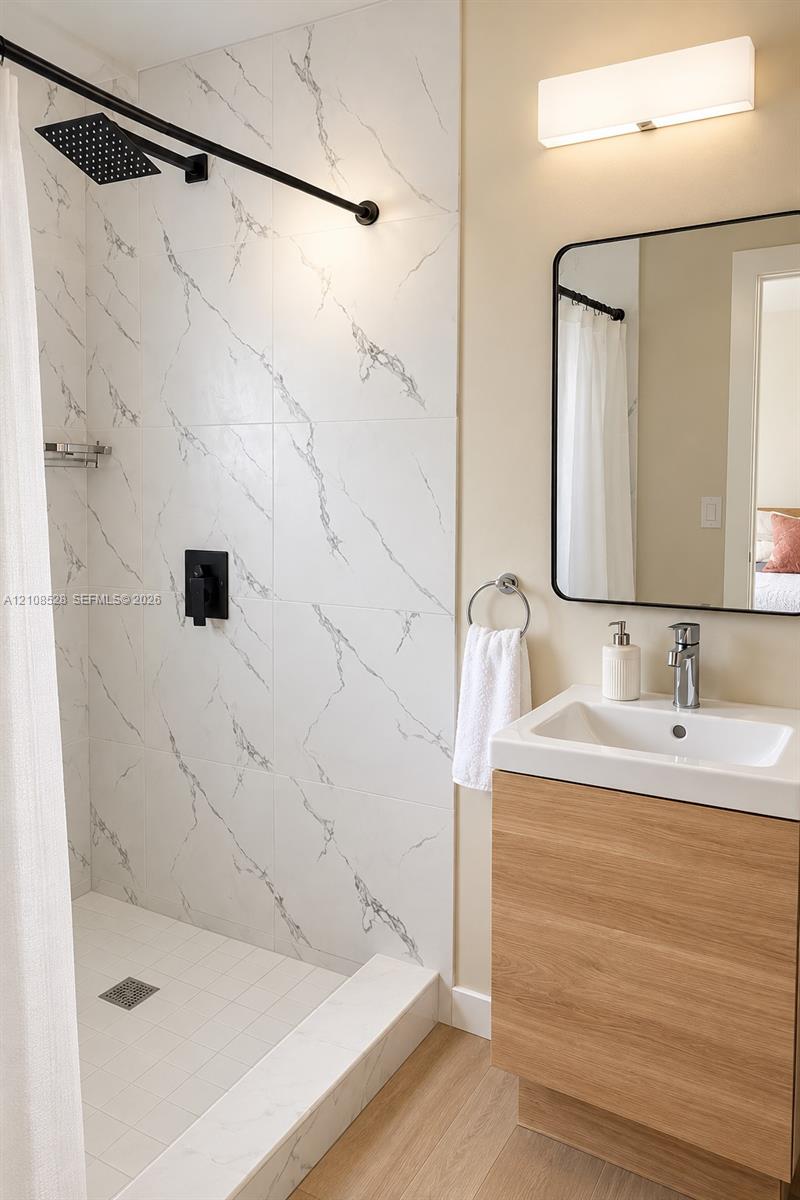 Walk-in shower featuring large-format tile with veining, matte black rainfall shower head, wood-finish vanity with integrated sink, rectangular wall mirror with black frame, and wall-mounted light fixture