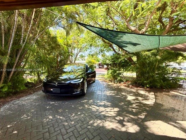 Herringbone-patterned paver driveway featuring mature tropical landscaping and a fixed overhead shade canopy