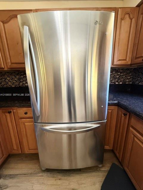 Stainless steel bottom-freezer refrigerator, dark-toned stone countertops, multi-colored mosaic tile backsplash, and wood-finish cabinetry Stainless steel bottom-freezer refrigerator, dark-toned stone countertops, multi-colored mosaic tile backsplash, and wood-finish cabinetry