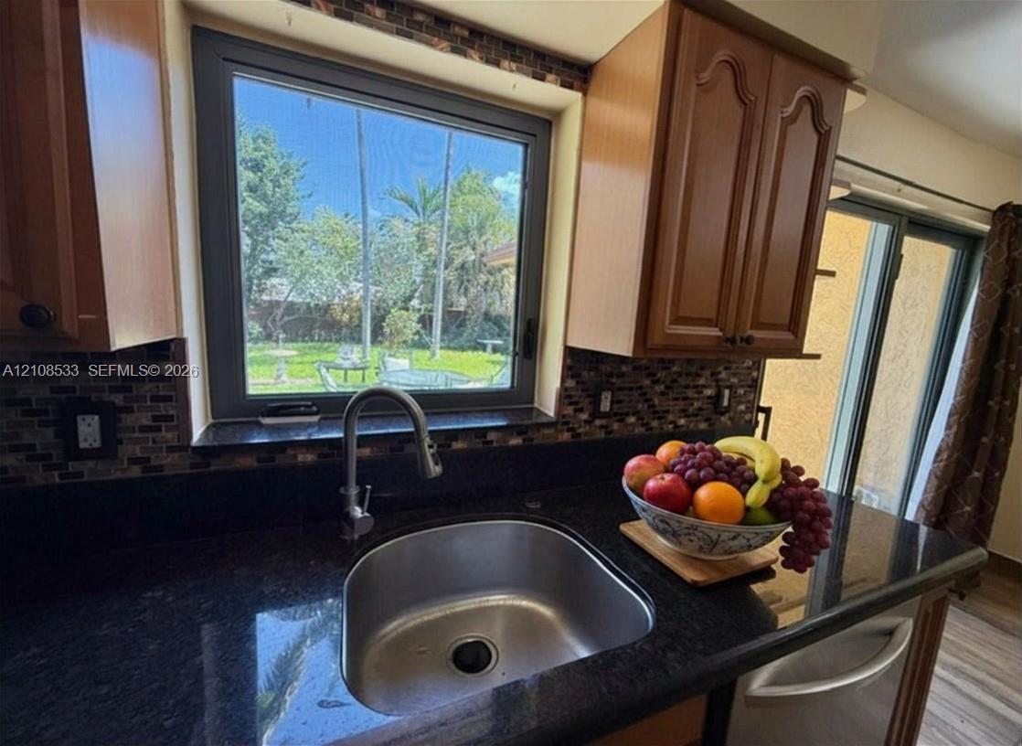 Kitchen featuring dark stone countertops, mosaic tile backsplash, wood-finish cabinetry, stainless steel undermount sink with high-arc faucet, and oversized window Kitchen featuring dark stone countertops, mosaic tile backsplash, wood-finish cabinetry, stainless steel undermount sink with high-arc faucet, and oversized window