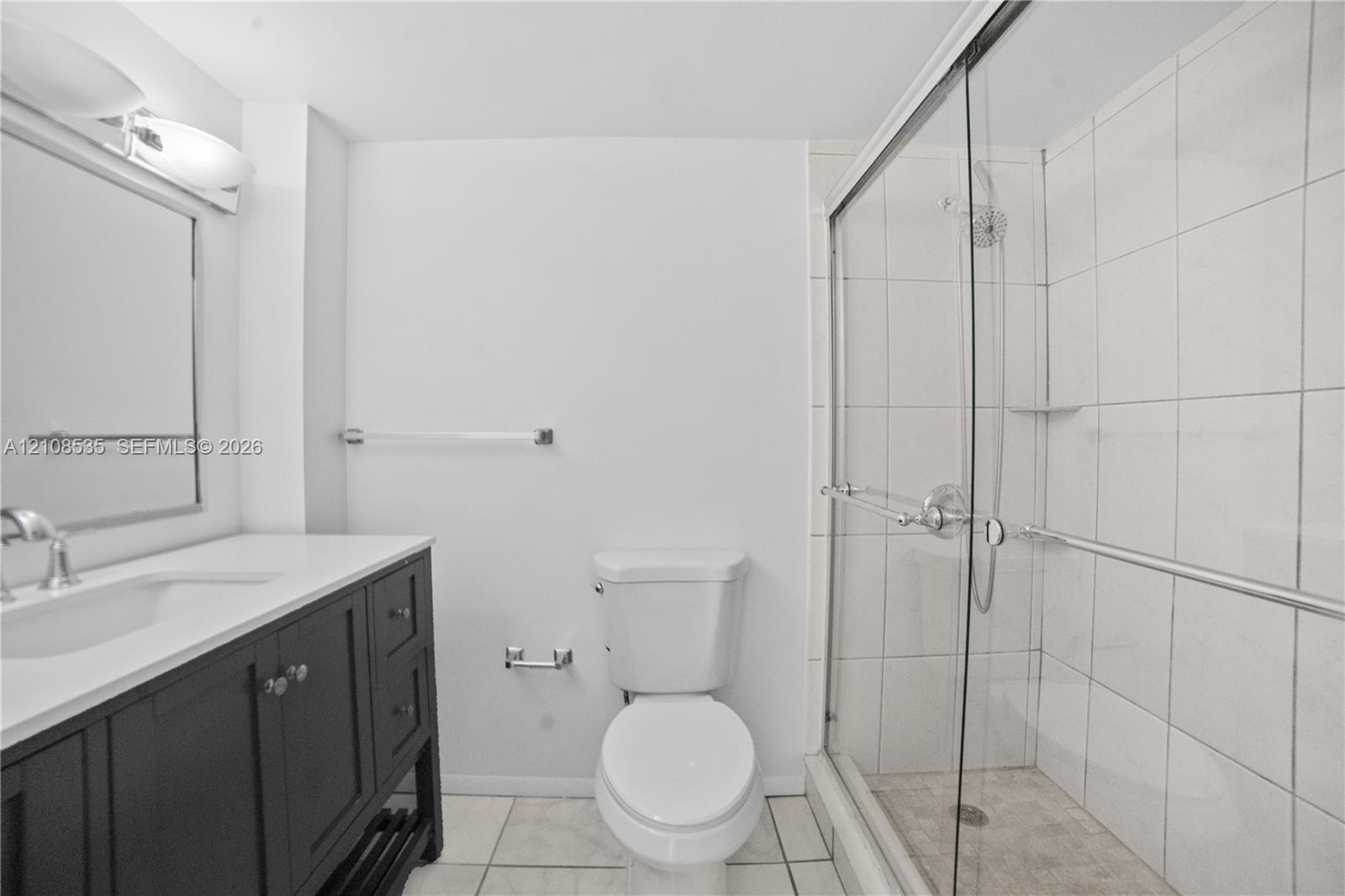 Dark wood vanity with a white countertop, integrated sink, walk-in shower with a sliding glass door, white wall tiling, and ceramic tile flooring