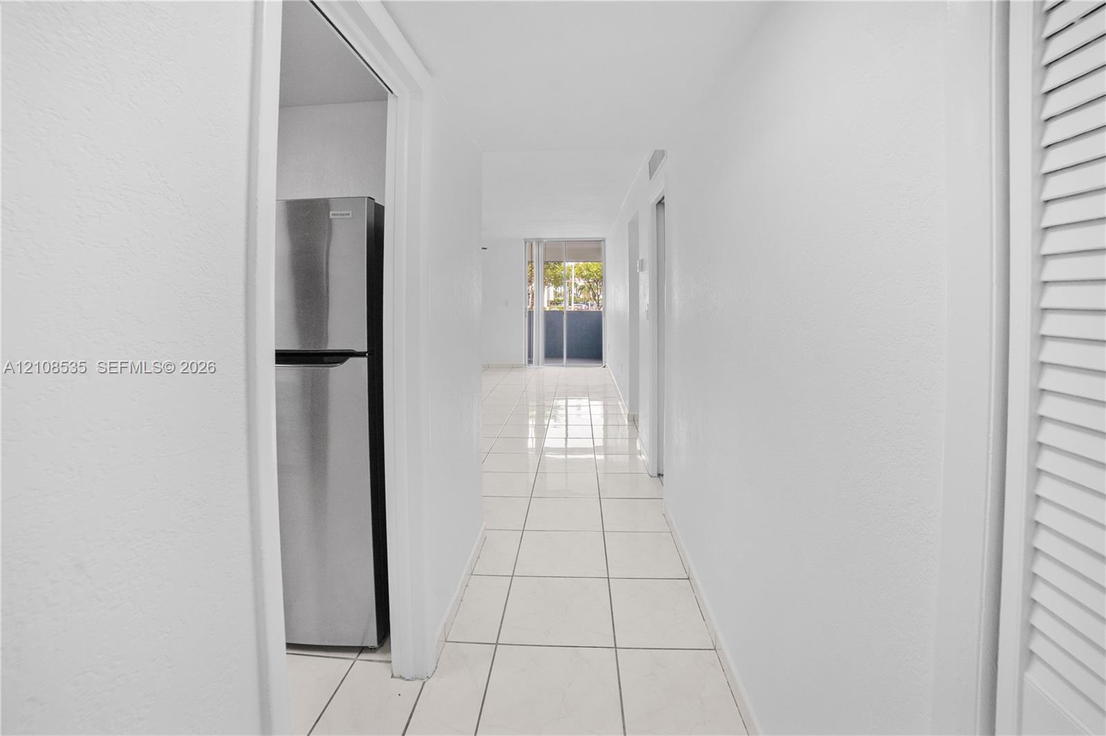 Hallway featuring neutral tile flooring, white walls, louvered closet doors, and a stainless steel refrigerator
