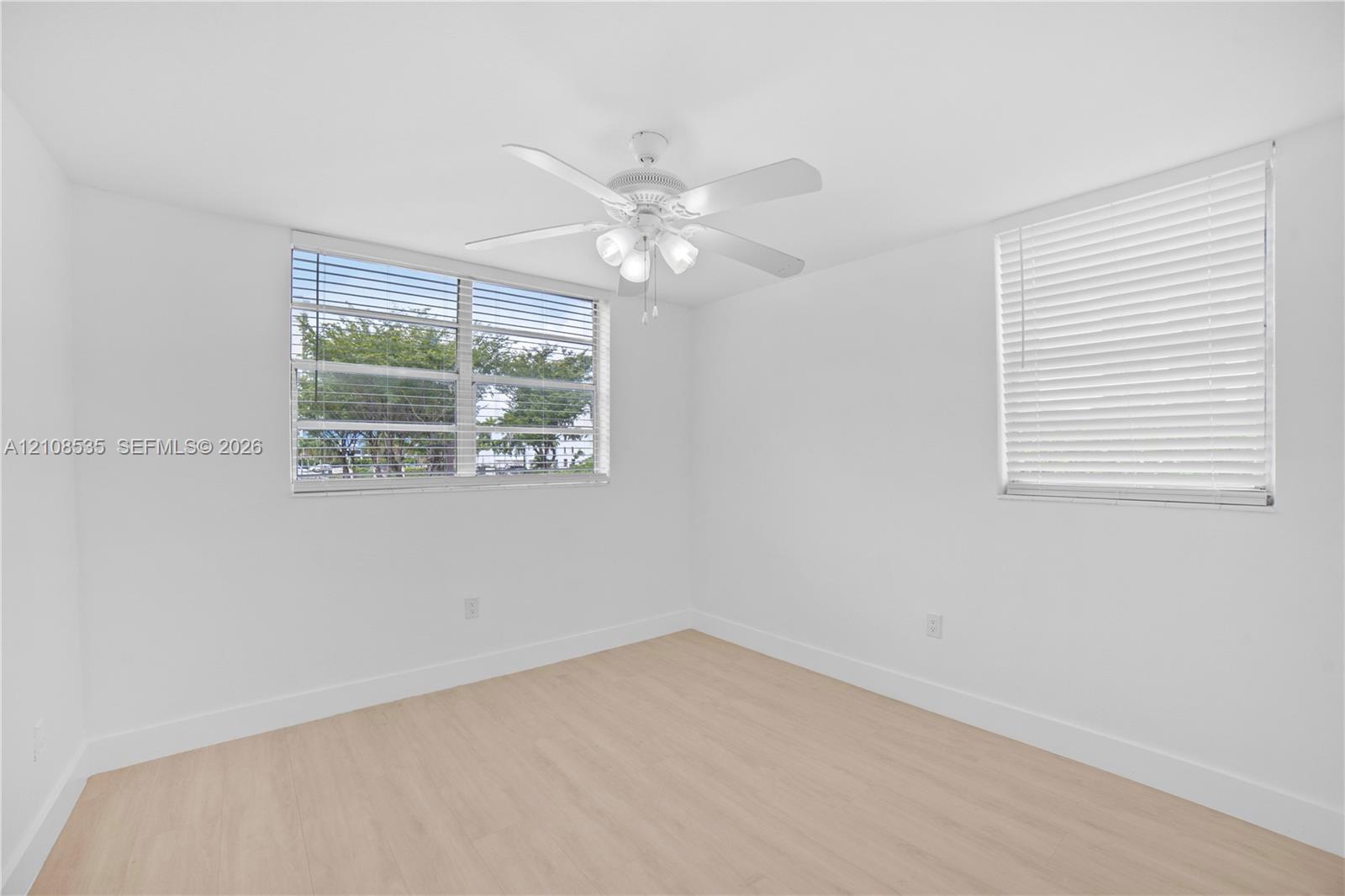 Neutral-toned interior space featuring wood-finish flooring, white walls, horizontal window blinds, and a white ceiling fan with integrated lighting