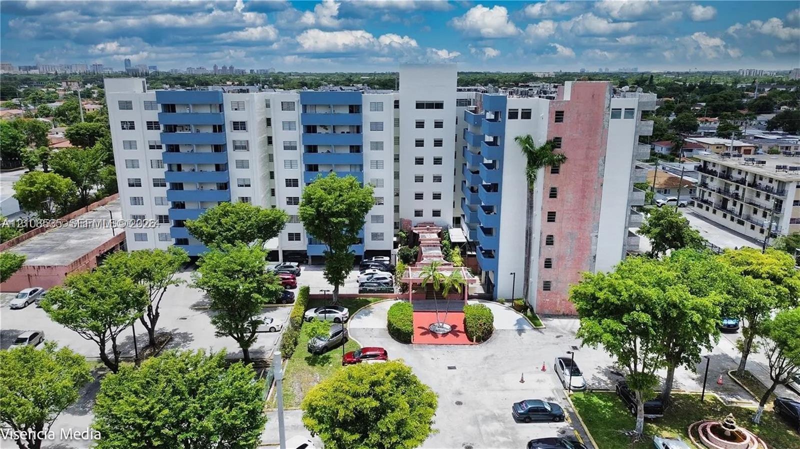 Multi-story building exterior with private balconies, surface parking, and landscaped grounds featuring mature trees and a decorative fountain