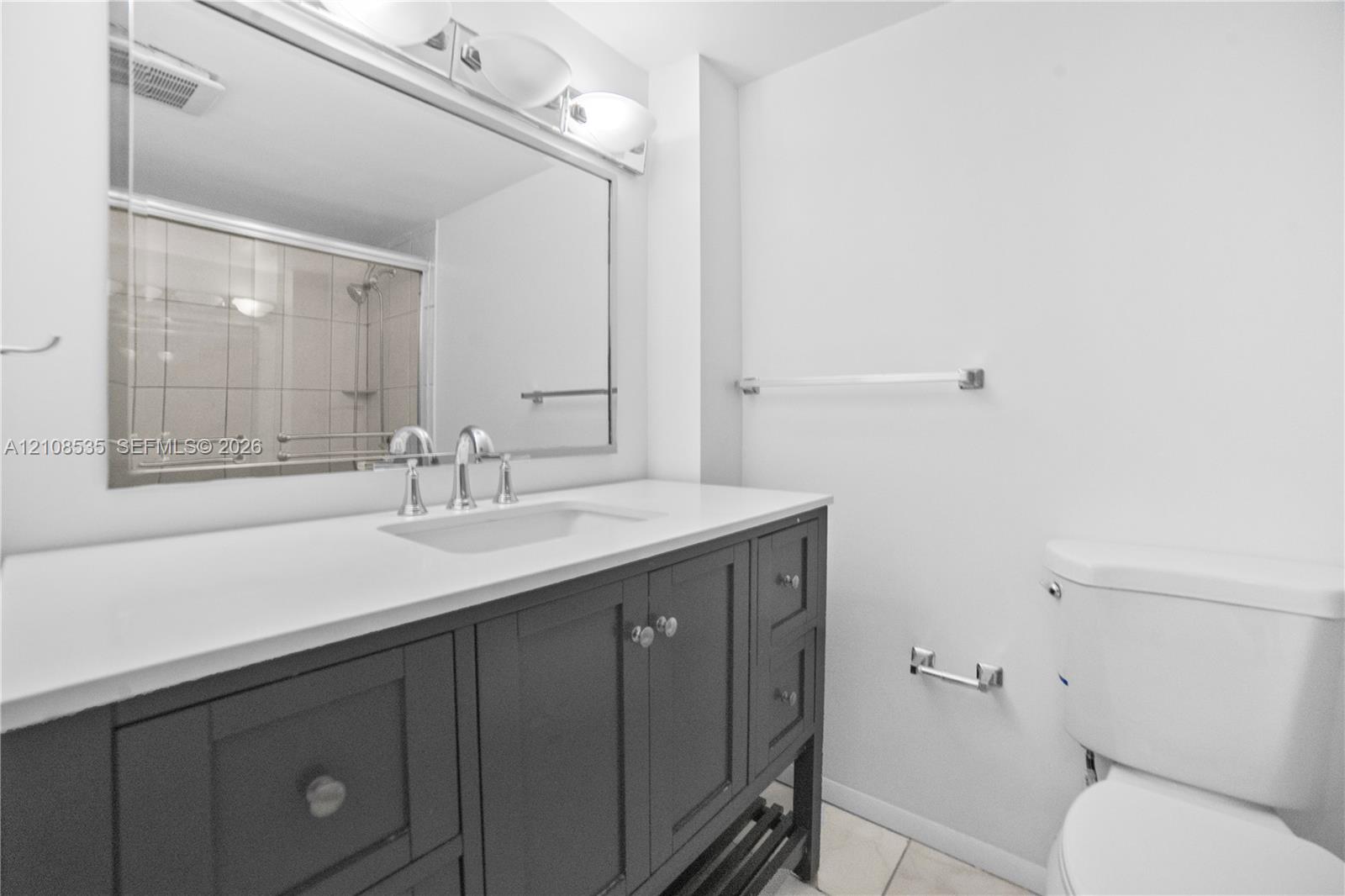 Charcoal-colored vanity with a light-toned solid surface countertop, undermount sink, polished chrome widespread faucet, large-format framed mirror, and integrated lighting