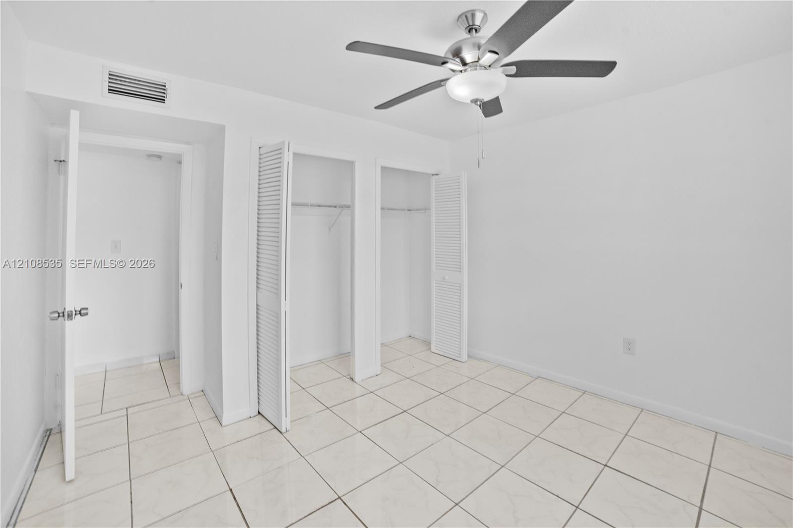 Room featuring light-toned tile flooring, neutral wall finishes, dual closets with louvered doors, and a ceiling-mounted light fixture with fan