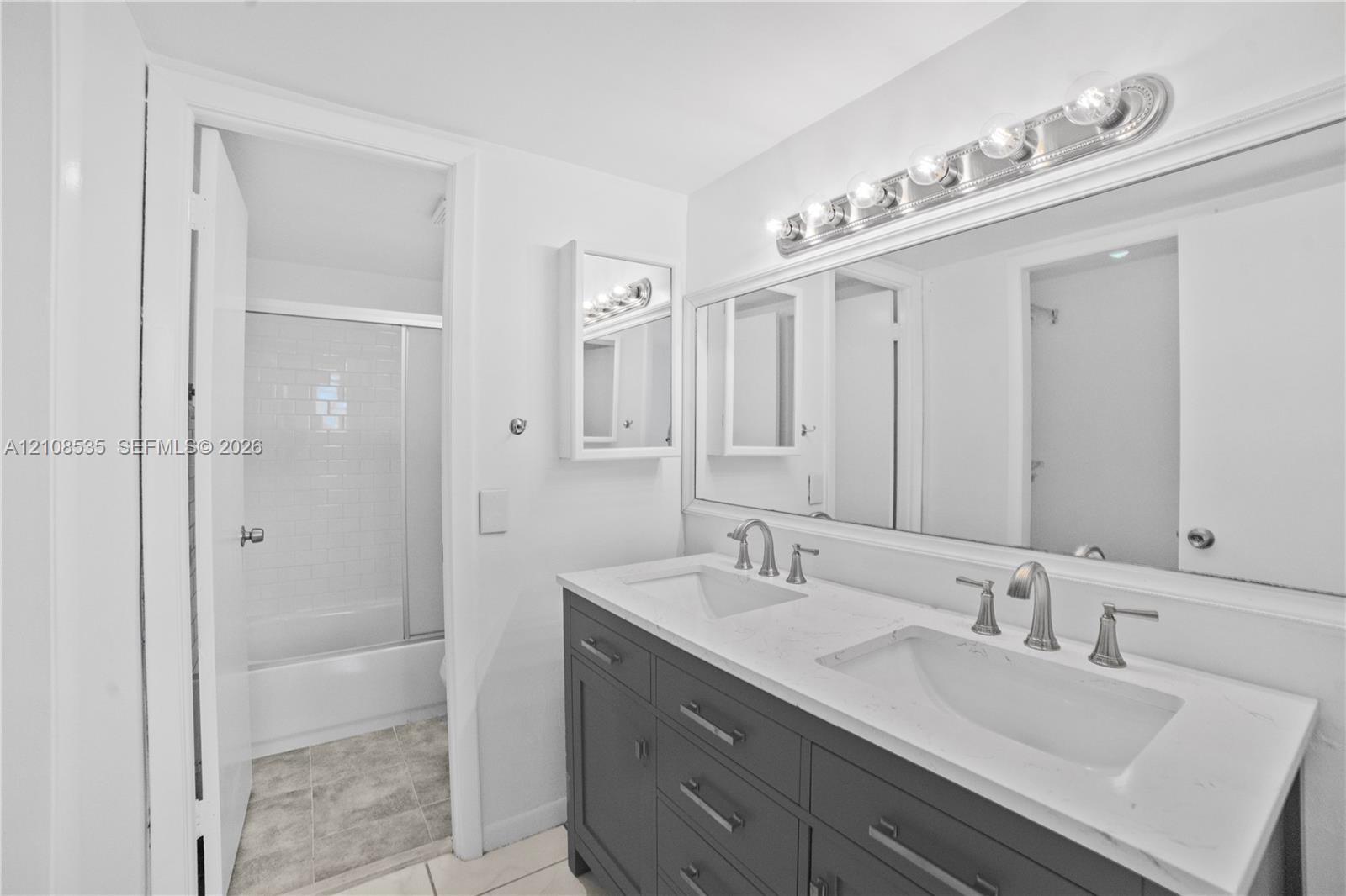 Dual-sink vanity with a solid-surface countertop, cabinetry featuring matte-finish hardware, large wall-mounted mirror, multi-bulb vanity lighting, and tub-shower combination with glass enclosure