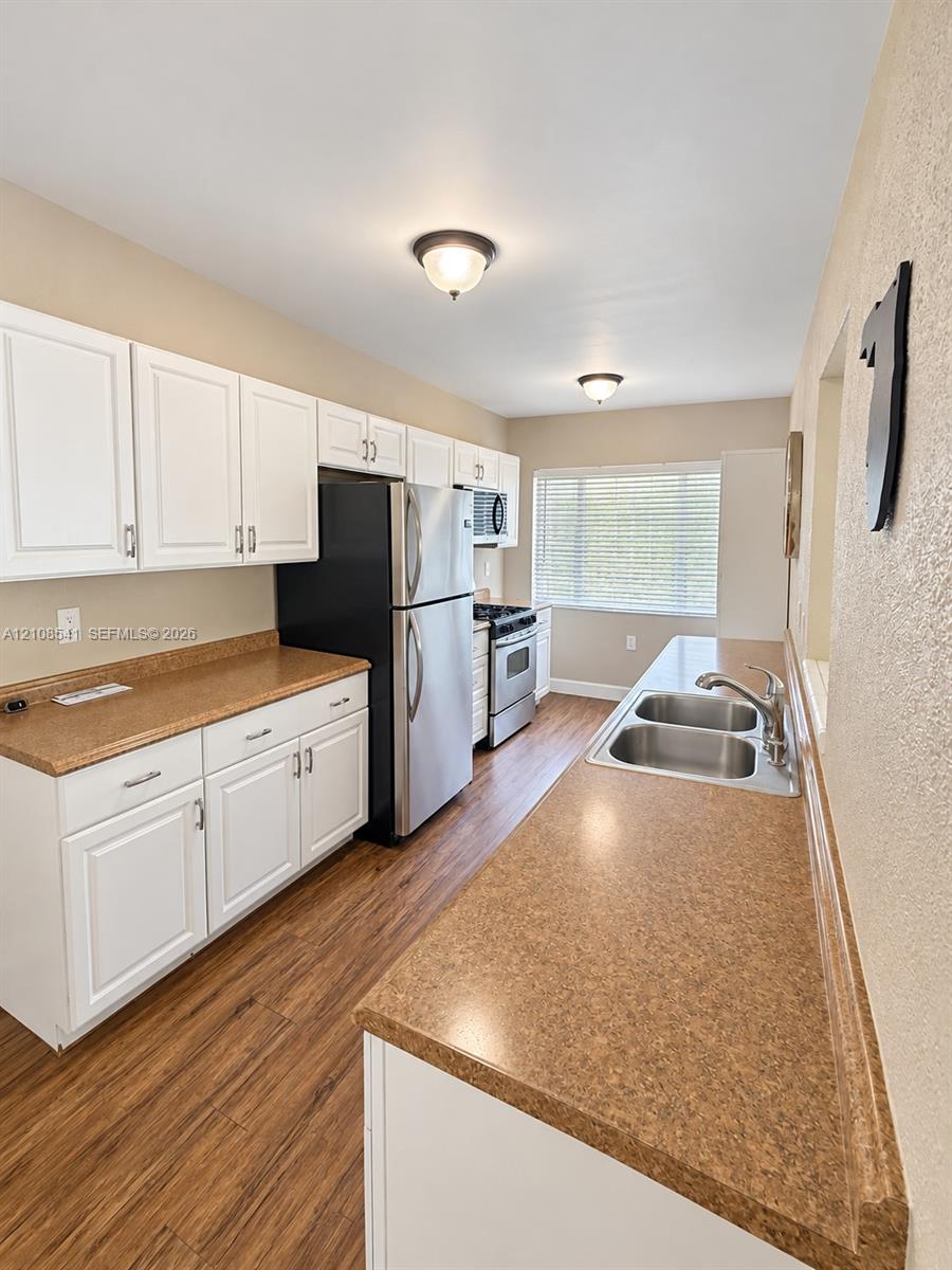 Kitchen featuring white shaker-style cabinetry, solid-surface countertops, wood-finish flooring, stainless steel appliances, and a dual-basin undermount sink Kitchen featuring white shaker-style cabinetry, solid-surface countertops, wood-finish flooring, stainless steel appliances, and a dual-basin undermount sink