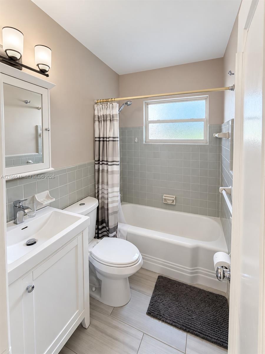 Bathroom featuring a white vanity cabinet with a integrated sink, light-toned tile flooring, light green wall tile surround, and a tub-shower combination with a frosted window Bathroom featuring a white vanity cabinet with a integrated sink, light-toned tile flooring, light green wall tile surround, and a tub-shower combination with a frosted window