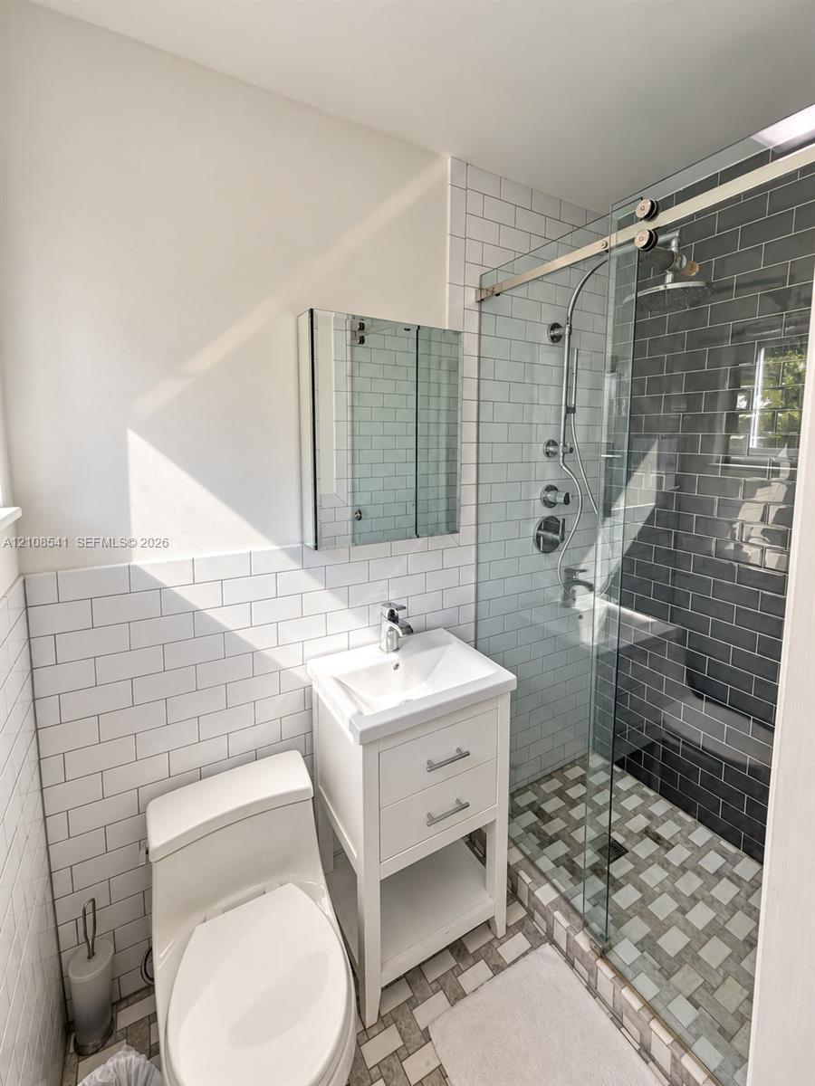 Bathroom featuring white subway tile wainscoting, a white pedestal-style vanity with metal hardware, a glass-enclosed shower with charcoal subway tile surround, multi-tone mosaic floor tiling, and a recessed mirrored medicine cabinet Bathroom featuring white subway tile wainscoting, a white pedestal-style vanity with metal hardware, a glass-enclosed shower with charcoal subway tile surround, multi-tone mosaic floor tiling, and a recessed mirrored medicine cabinet