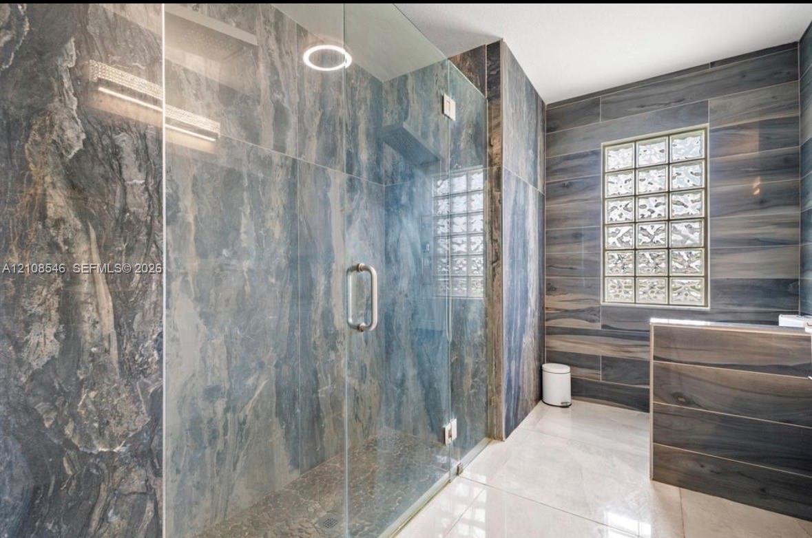 Walk-in shower enclosure with glass panels, large-format stone-finish tile surround, glass block window, and light-tone tile flooring