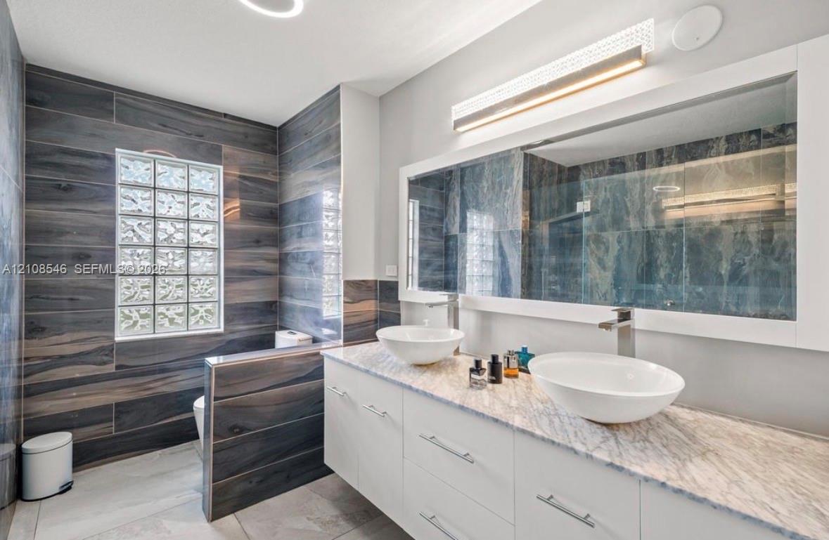 Bathroom featuring a dual-sink vanity with a stone countertop, wall-to-wall mirror, glass block window, and floor-to-ceiling wood-finish tile wall
