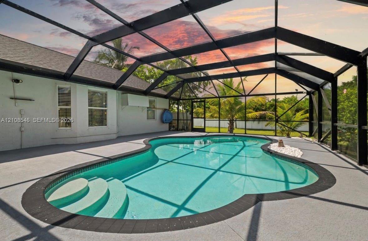 Screen-enclosed pool area featuring a freeform swimming pool, integrated shallow-water entry steps, textured concrete decking, and a dark metal frame structure Screen-enclosed pool area featuring a freeform swimming pool, integrated shallow-water entry steps, textured concrete decking, and a dark metal frame structure