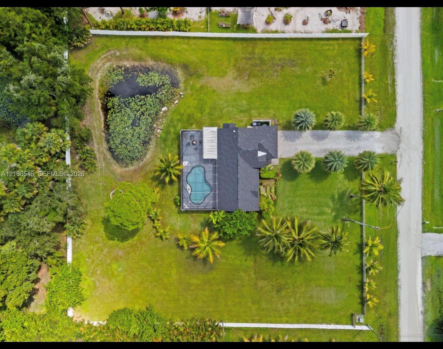 Aerial perspective of a property featuring a screened-in pool, a private pond, expansive lawn, mature palm trees, and perimeter fencing