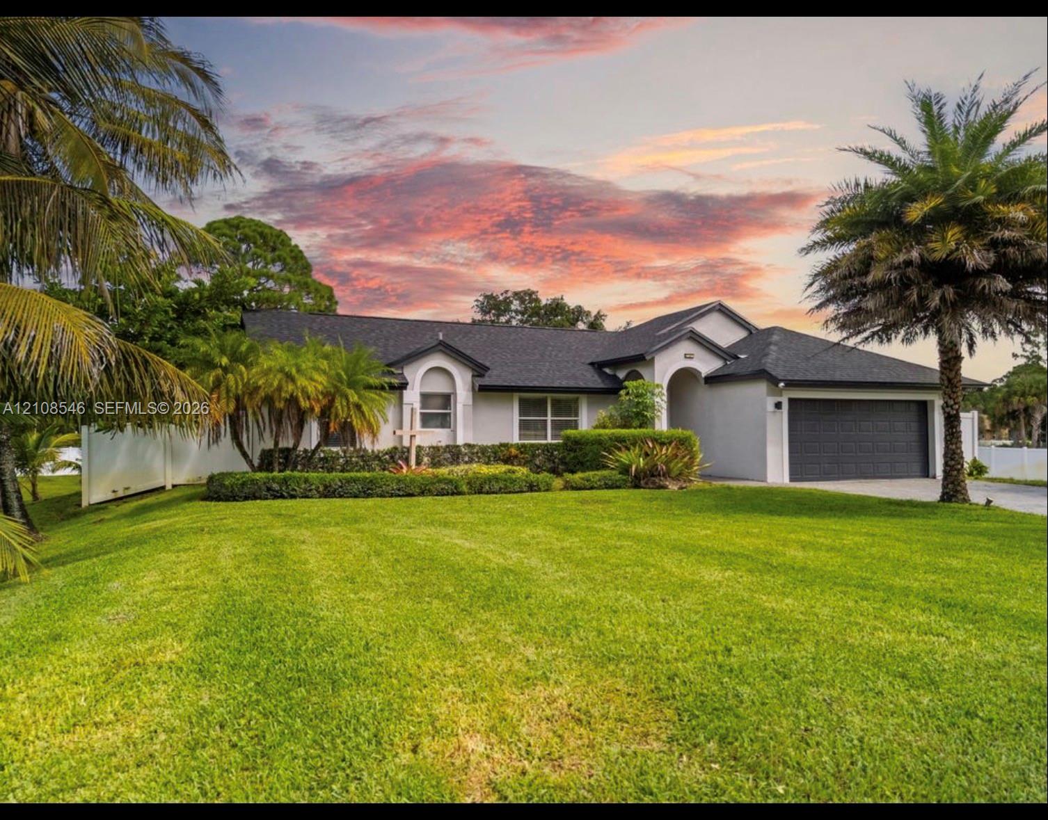 Single-story exterior featuring a light-toned stucco faade, a dark shingle roof, an attached two-car garage, manicured lawn, and mature palm landscaping Single-story exterior featuring a light-toned stucco faade, a dark shingle roof, an attached two-car garage, manicured lawn, and mature palm landscaping