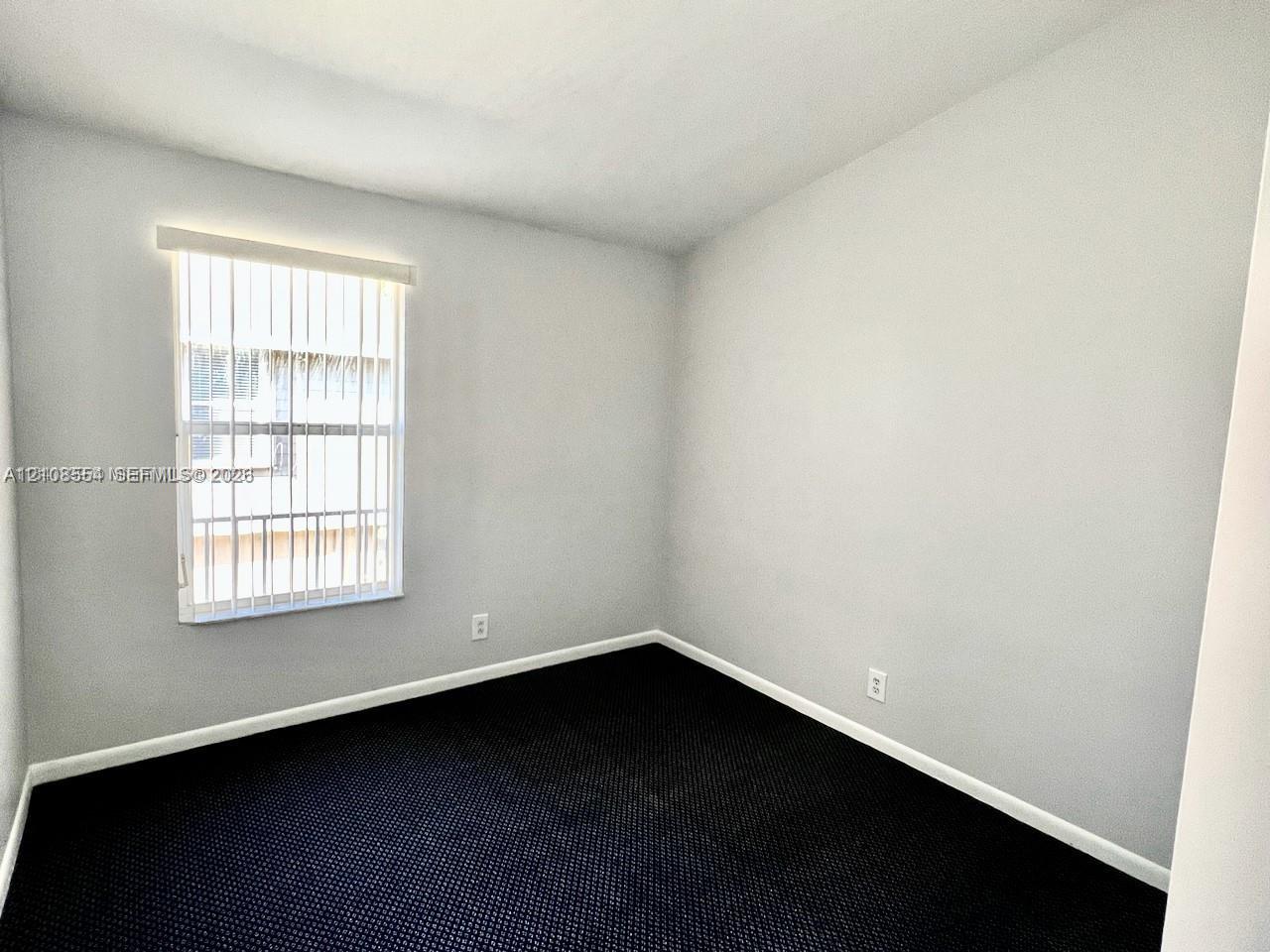 Neutral-toned interior space featuring dark wall-to-wall carpeting, light-painted walls, and a single-pane window with vertical blinds