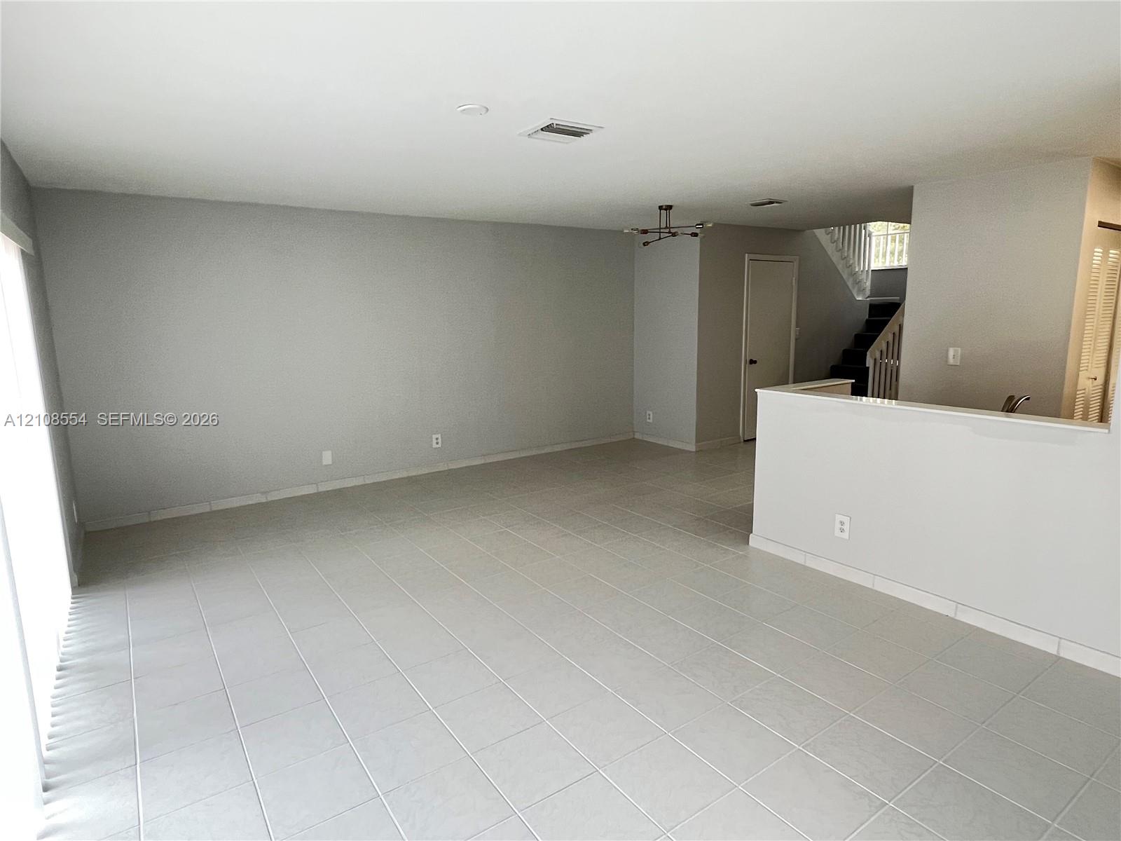 Neutral interior space featuring large-format ceramic tile flooring, a half-wall partition, a multi-arm modern light fixture, and a staircase with vertical wood balusters