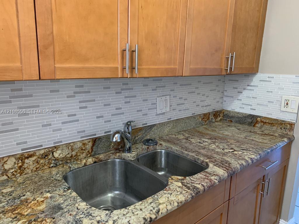 Natural stone countertop with an integrated double-basin stainless steel sink, polished chrome faucet, linear mosaic tile backsplash, and light-tone wood-finish cabinetry featuring brushed metal hardware