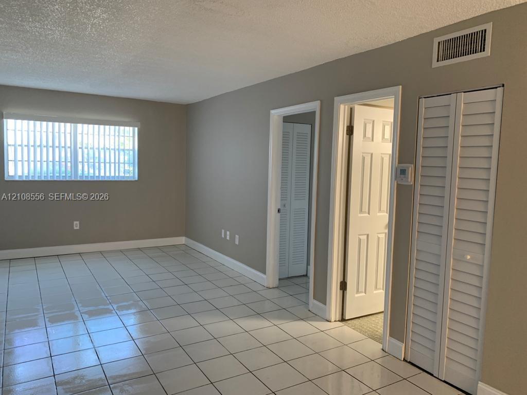 Neutral-toned interior space featuring light-colored ceramic tile flooring, a large window with vertical blinds, and dual white louvered closet doors Neutral-toned interior space featuring light-colored ceramic tile flooring, a large window with vertical blinds, and dual white louvered closet doors