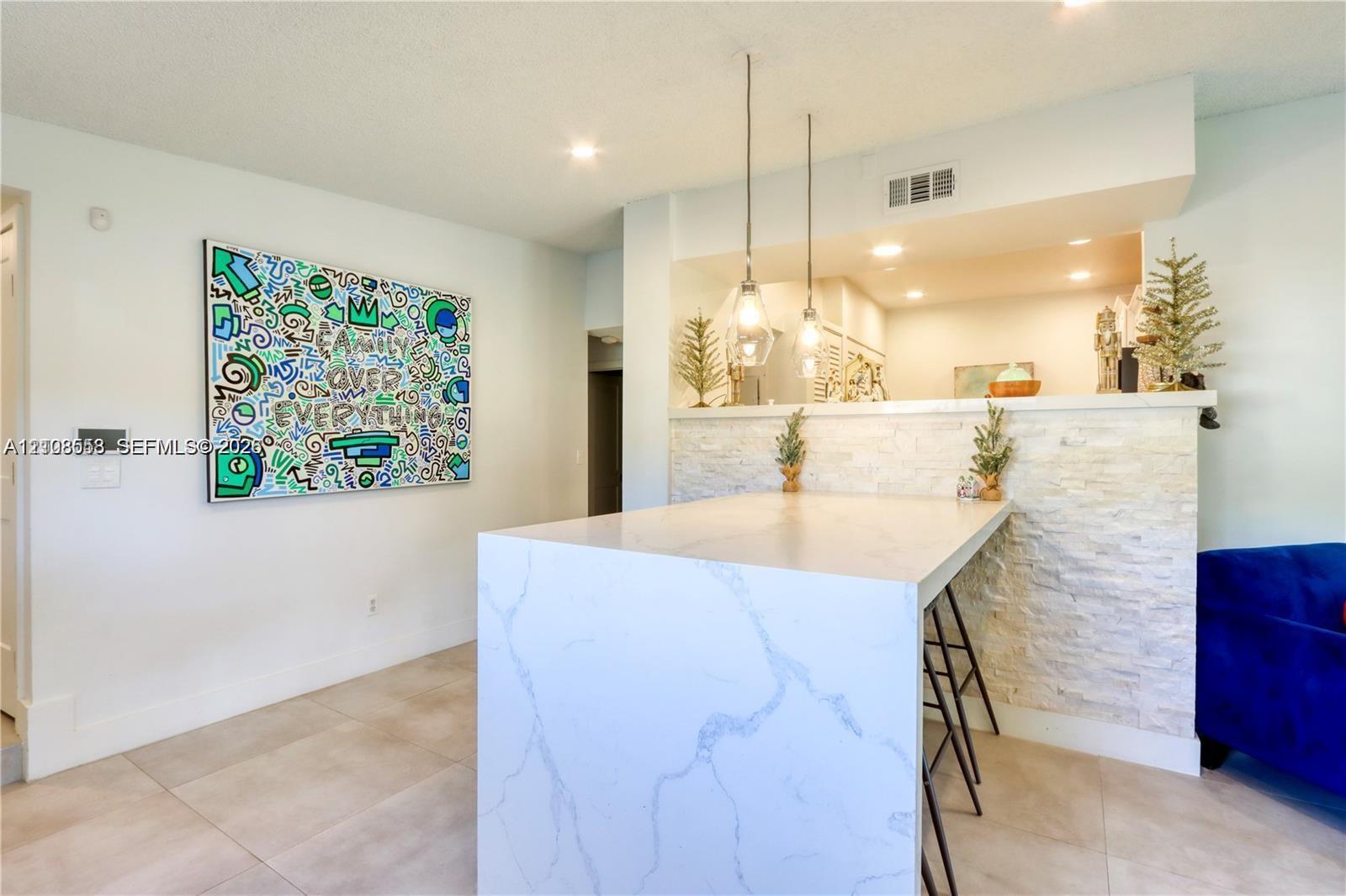 Kitchen island featuring a stone-finish waterfall countertop, stacked stone facade, dual pendant lighting, and neutral tile flooring