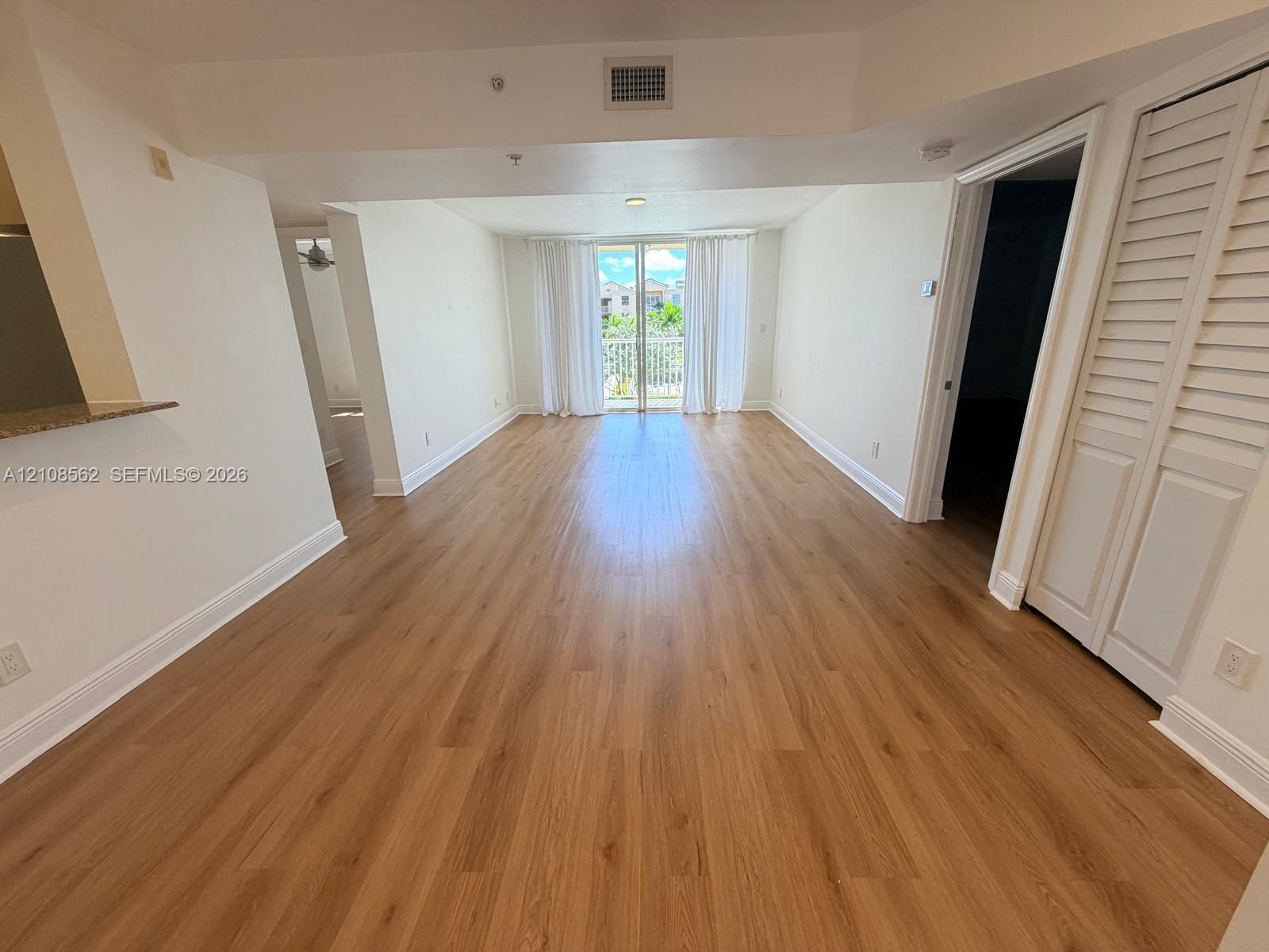 Open-concept interior featuring wood-finish flooring, louvered closet doors, and a glass-paneled sliding door opening to a balcony Open-concept interior featuring wood-finish flooring, louvered closet doors, and a glass-paneled sliding door opening to a balcony