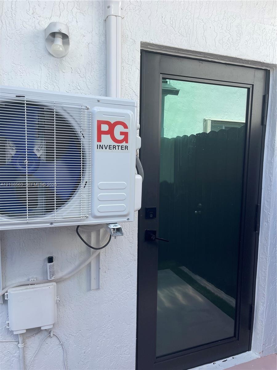 Exterior wall-mounted ductless inverter AC unit, black-framed glass entry door, and wall-mounted exterior light fixture