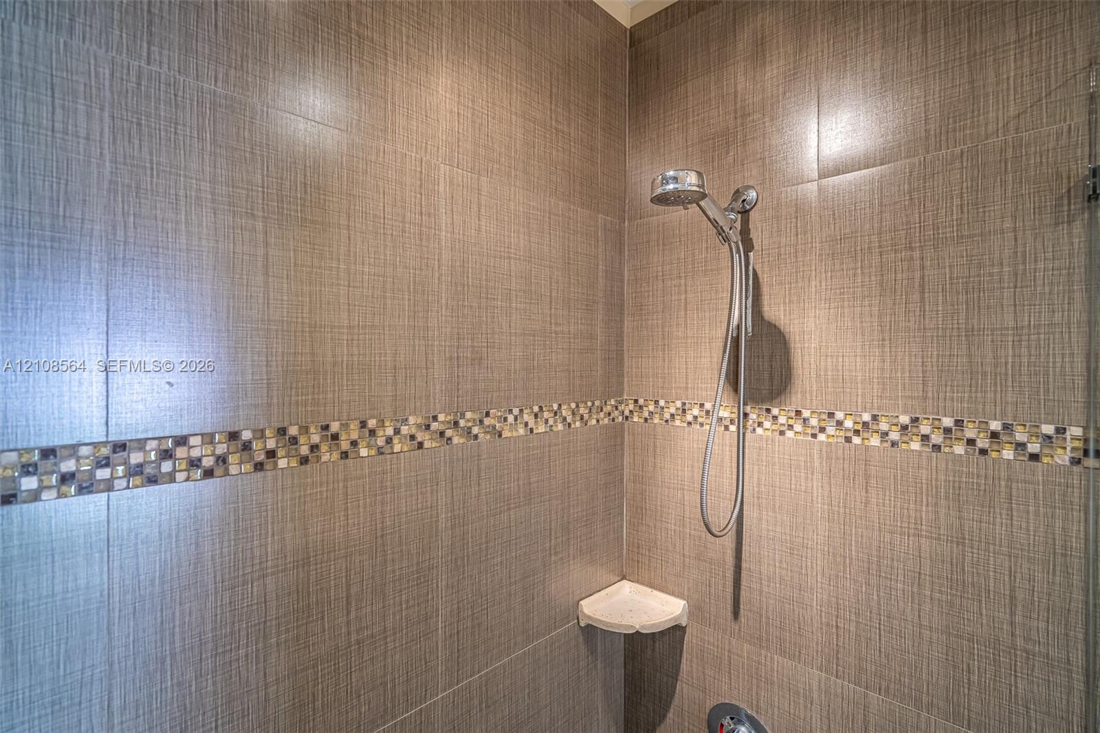 Shower enclosure featuring large-format textured wall tile, decorative mosaic tile inlay, chrome handheld shower fixture, and a built-in corner shelf