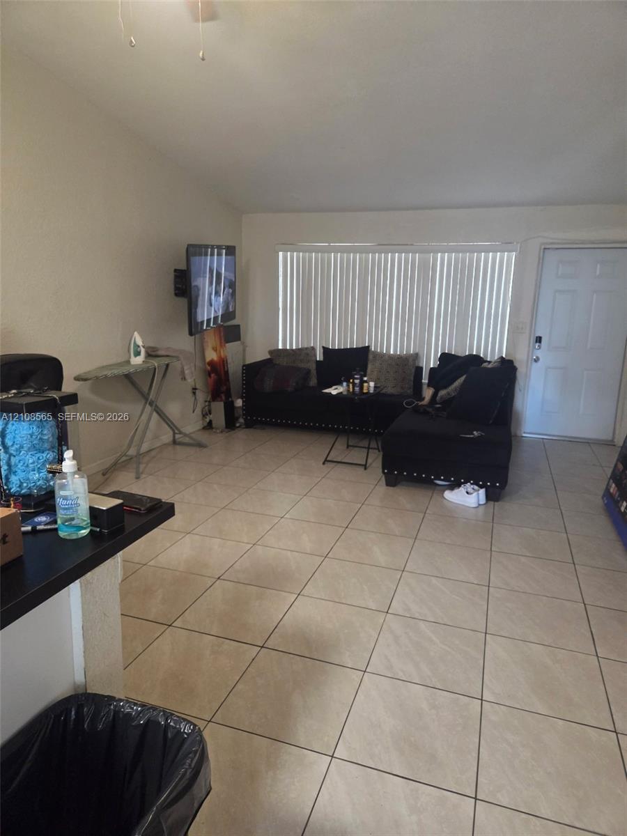 Open-concept room featuring light-toned tile flooring, vaulted ceiling, and large window with vertical blinds