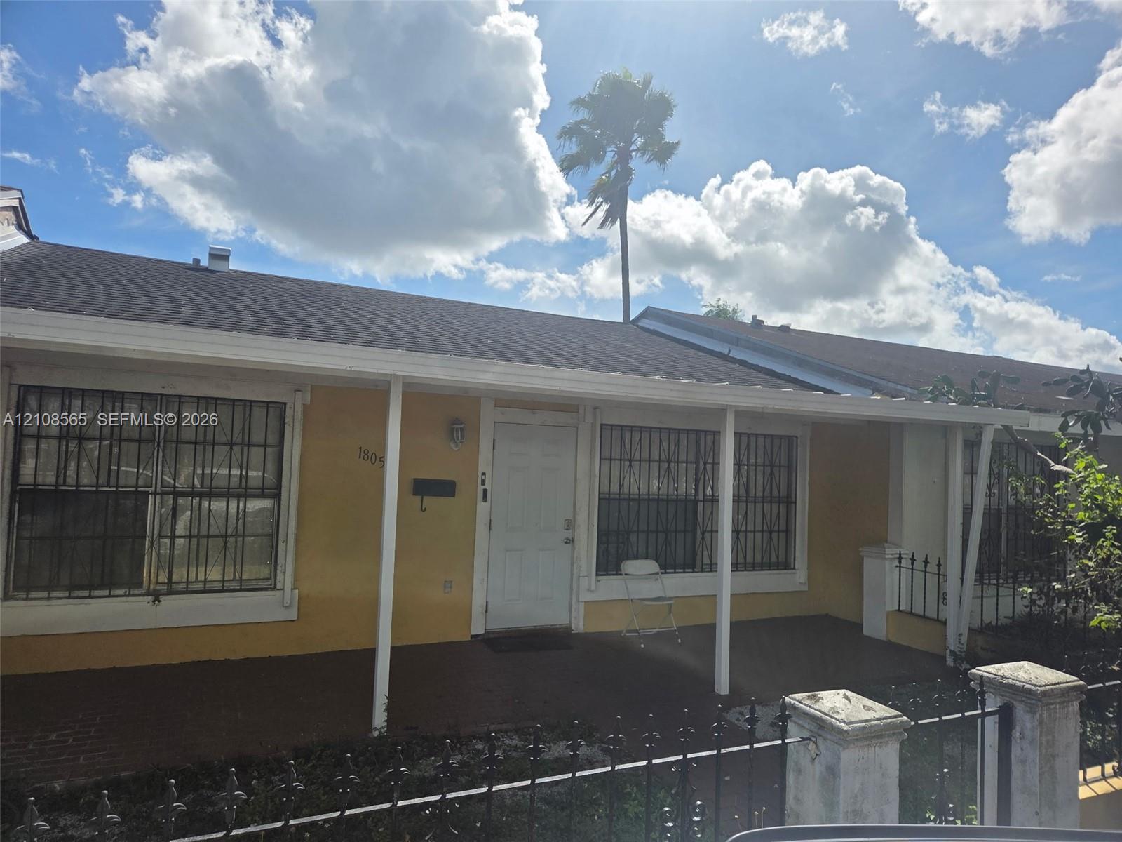 Single-story residence featuring a covered front porch, exterior metal window security bars, a concrete front patio, and a perimeter metal fence with masonry pillars Single-story residence featuring a covered front porch, exterior metal window security bars, a concrete front patio, and a perimeter metal fence with masonry pillars