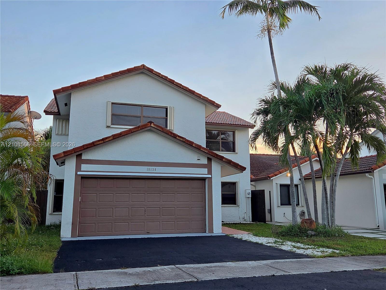 Two-story exterior featuring a light-toned stucco faade, terracotta-style roof tiles, an attached garage with a sectional door, and mature palm trees