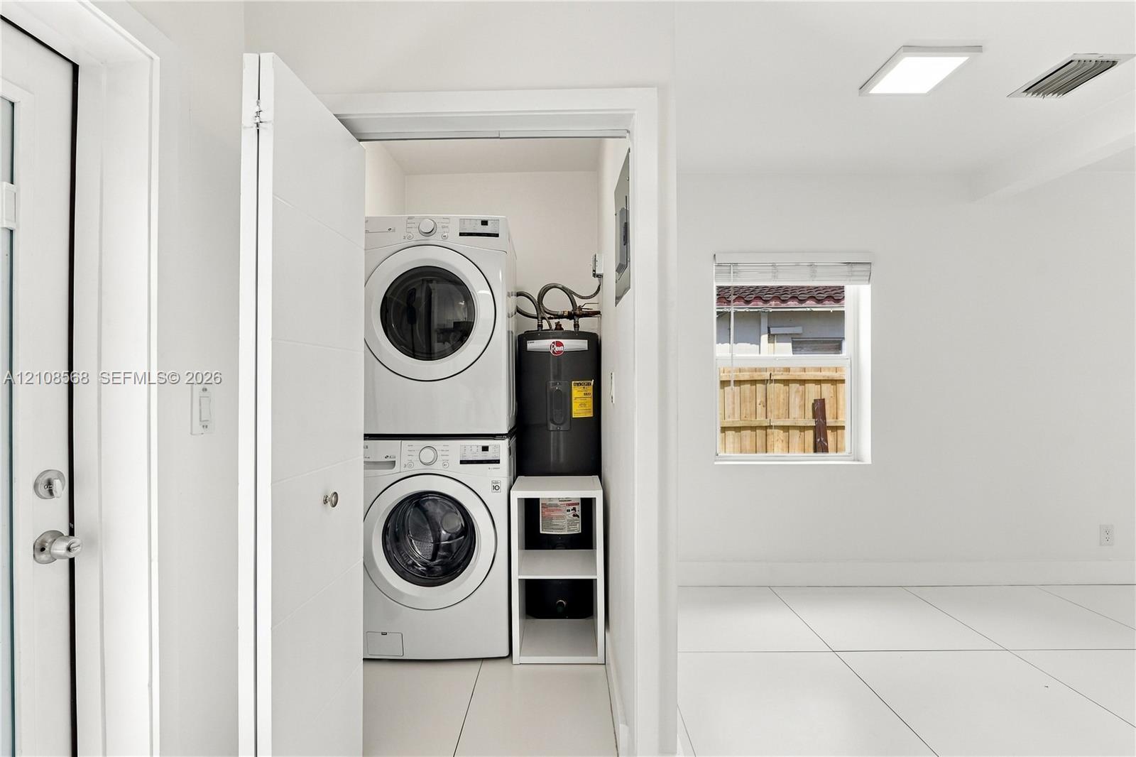 Utility closet featuring a stacked front-loading washer and dryer, water heater, white tile flooring, and flush-mount LED lighting