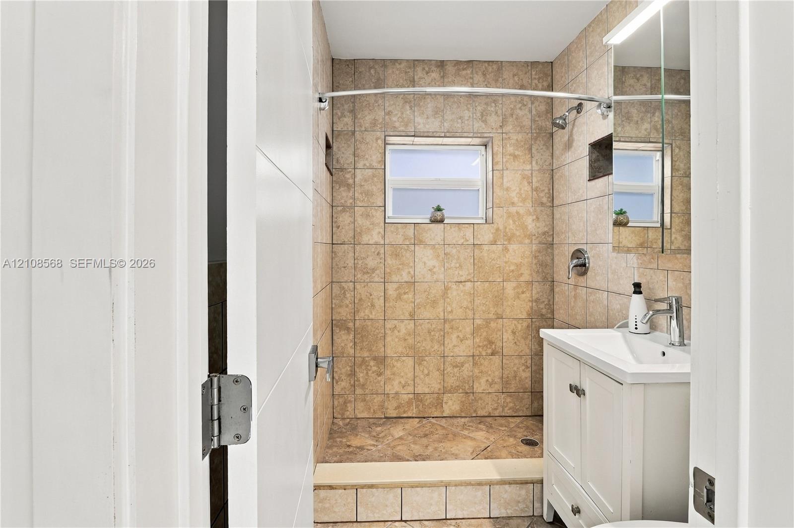 Bathroom featuring a walk-in shower with stone-finish tile surround, a built-in shower niche, a white vanity with a rectangular integrated sink, and a mirrored medicine cabinet