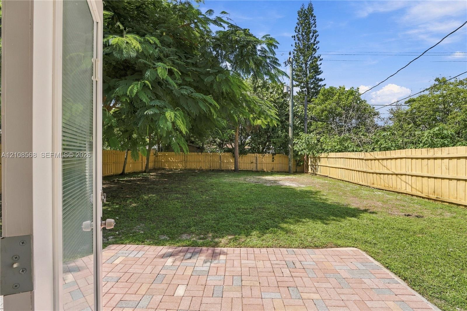 Backyard featuring a paved brick patio, manicured lawn, perimeter wood fencing, and mature trees