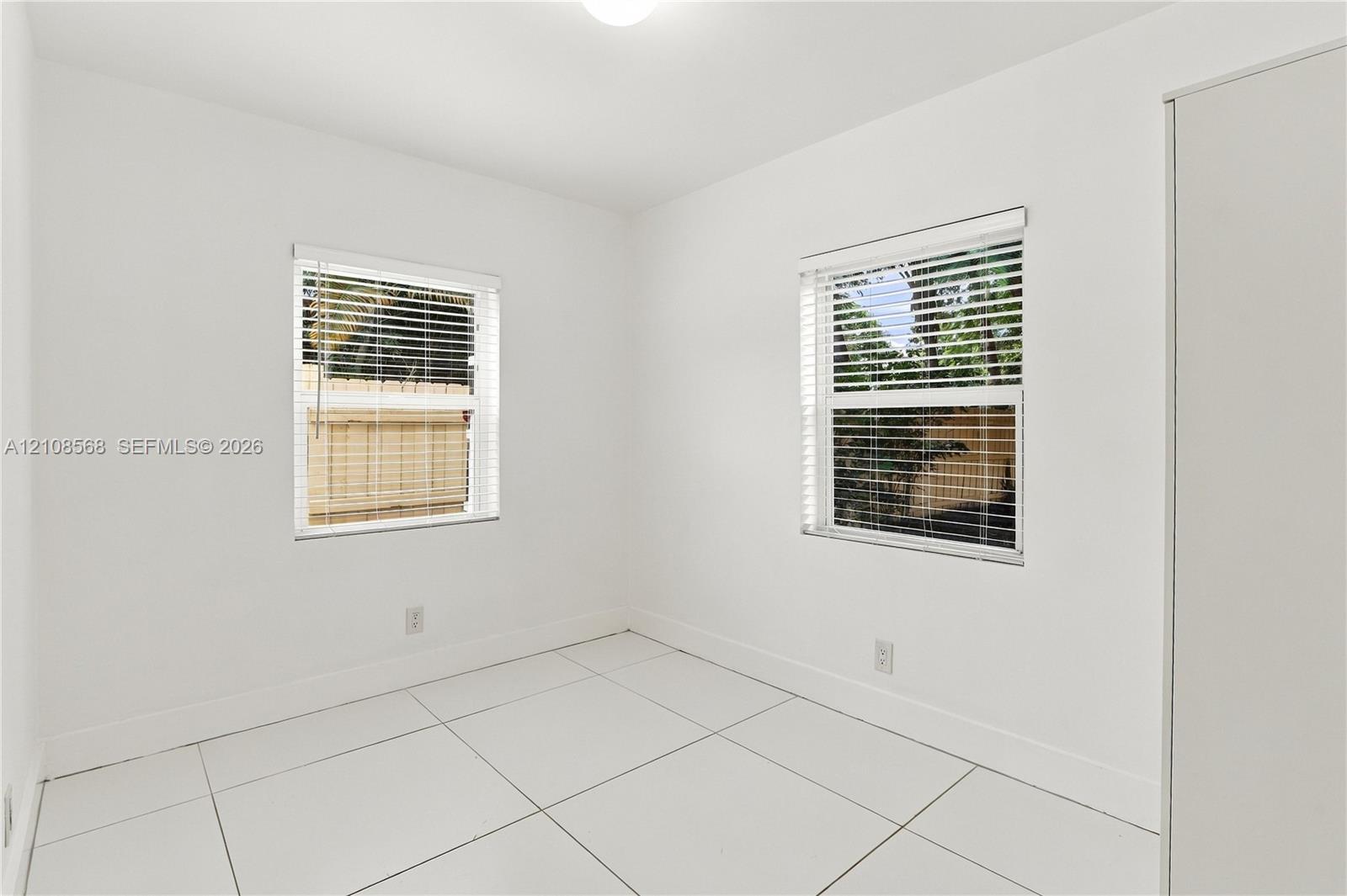 Bright room featuring large-format white tile flooring, white walls, dual windows with fitted blinds, and a full-height built-in wardrobe