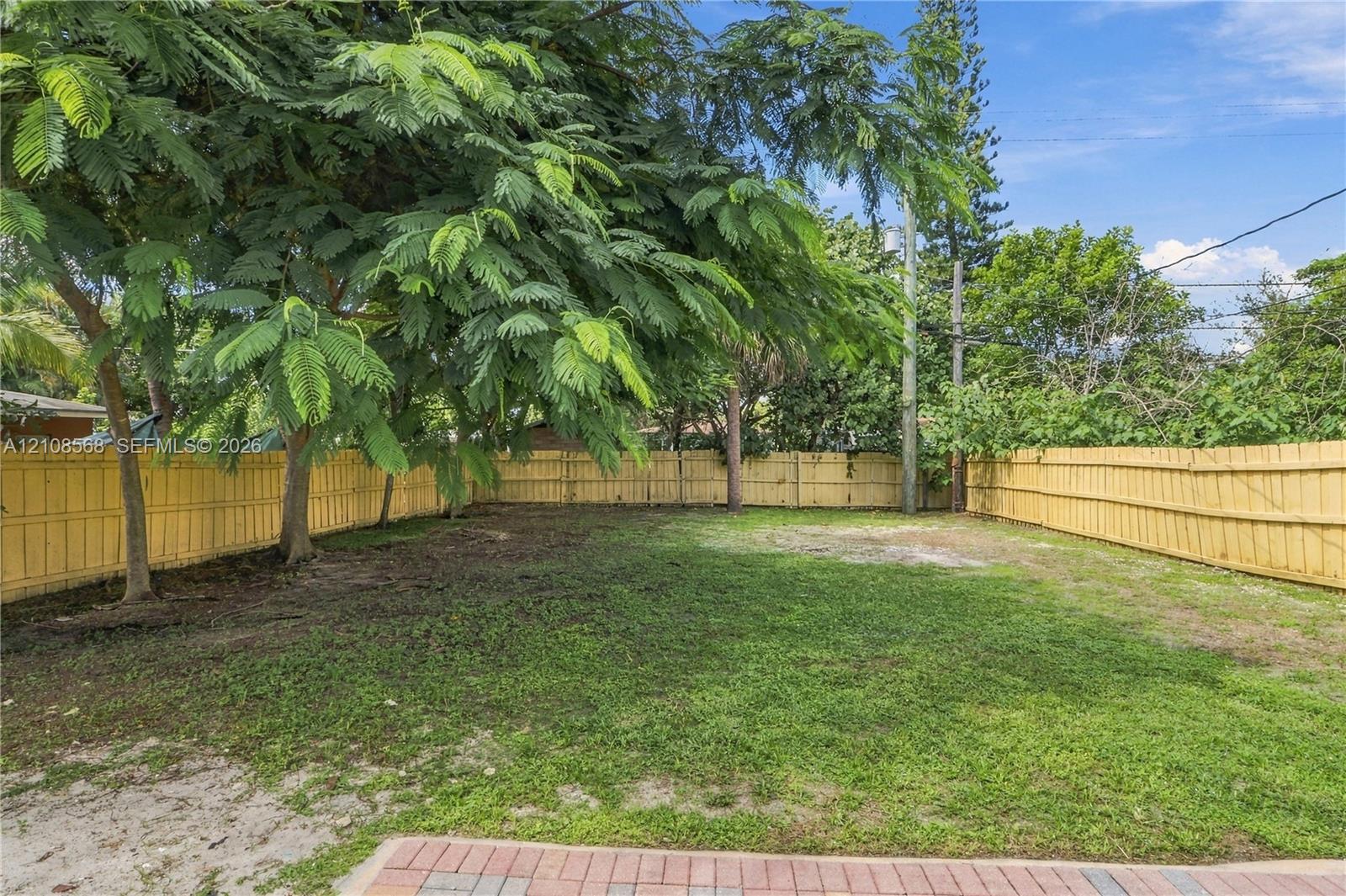 Rear yard featuring a grass lawn, perimeter wood fencing, mature trees, and a paved brick patio
