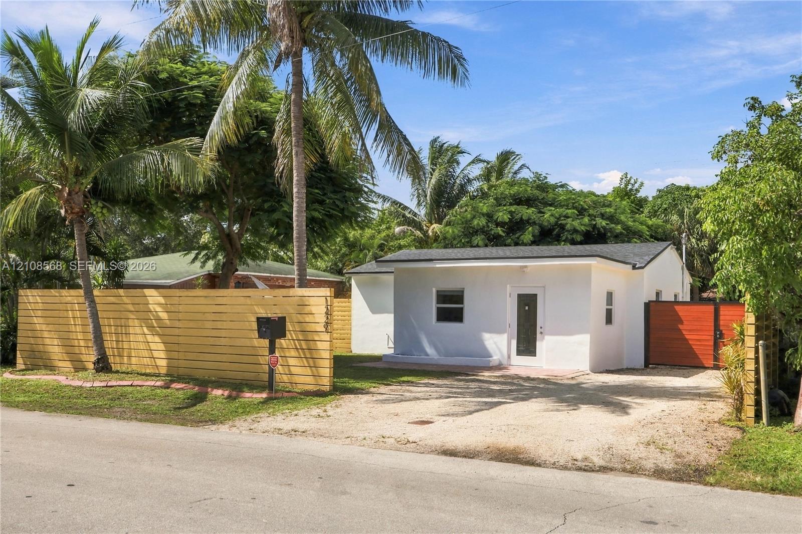 Single-story exterior featuring white stucco siding, a shingled roof, a horizontal wood privacy fence, a gravel driveway, and a gated side entrance