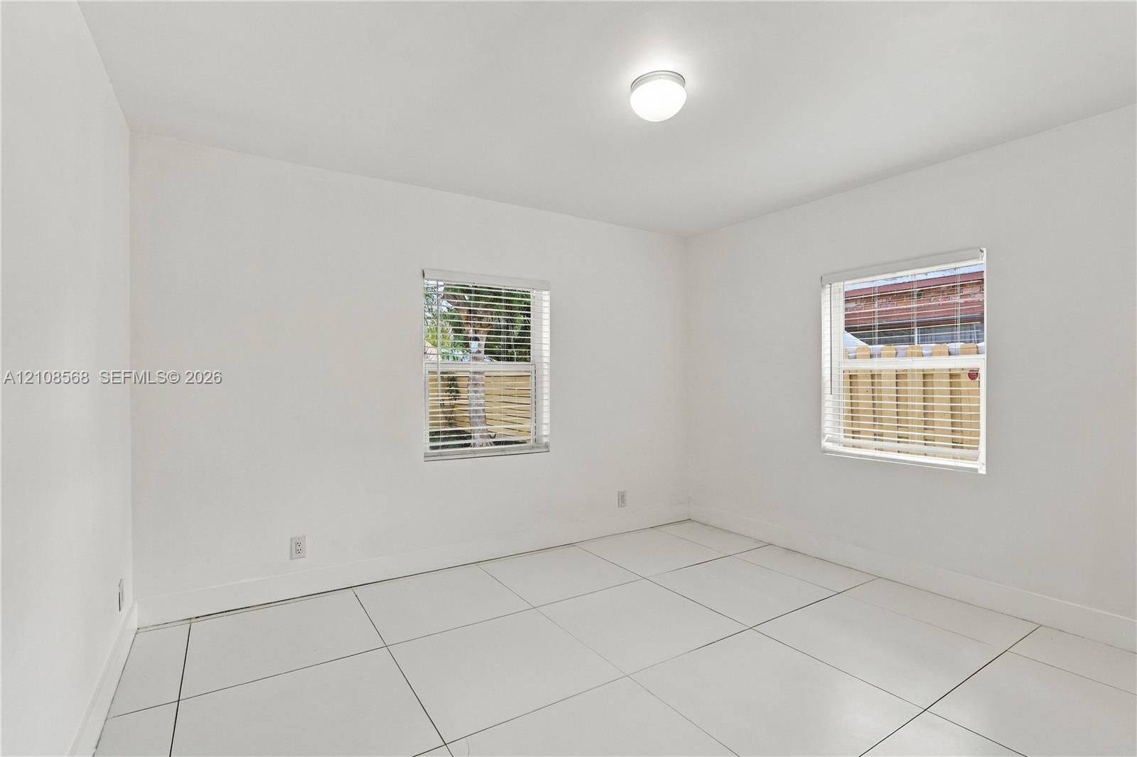 Neutral-toned room featuring large-format tile flooring, bright interior walls, two windows with horizontal blinds, and a flush-mount ceiling light fixture