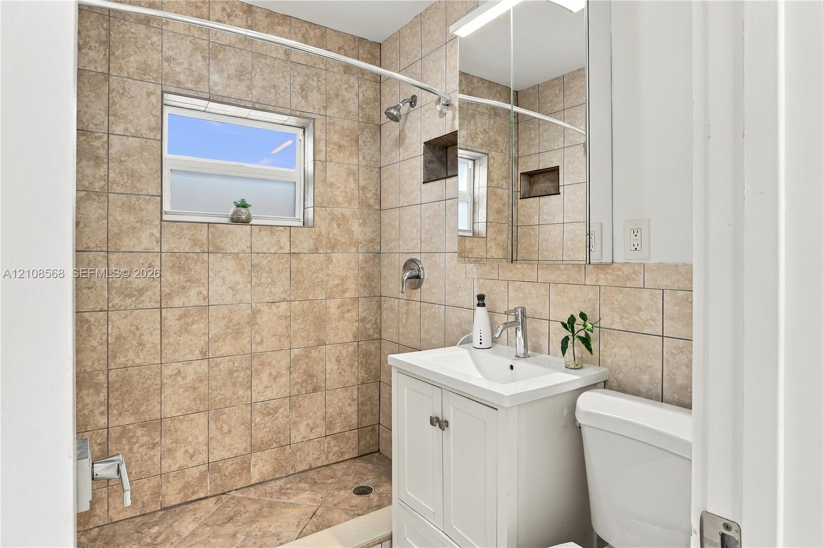 Bathroom featuring tile-surround walk-in shower with inset shelving, white vanity with integrated sink, chrome-finish faucet, and mirrored medicine cabinet