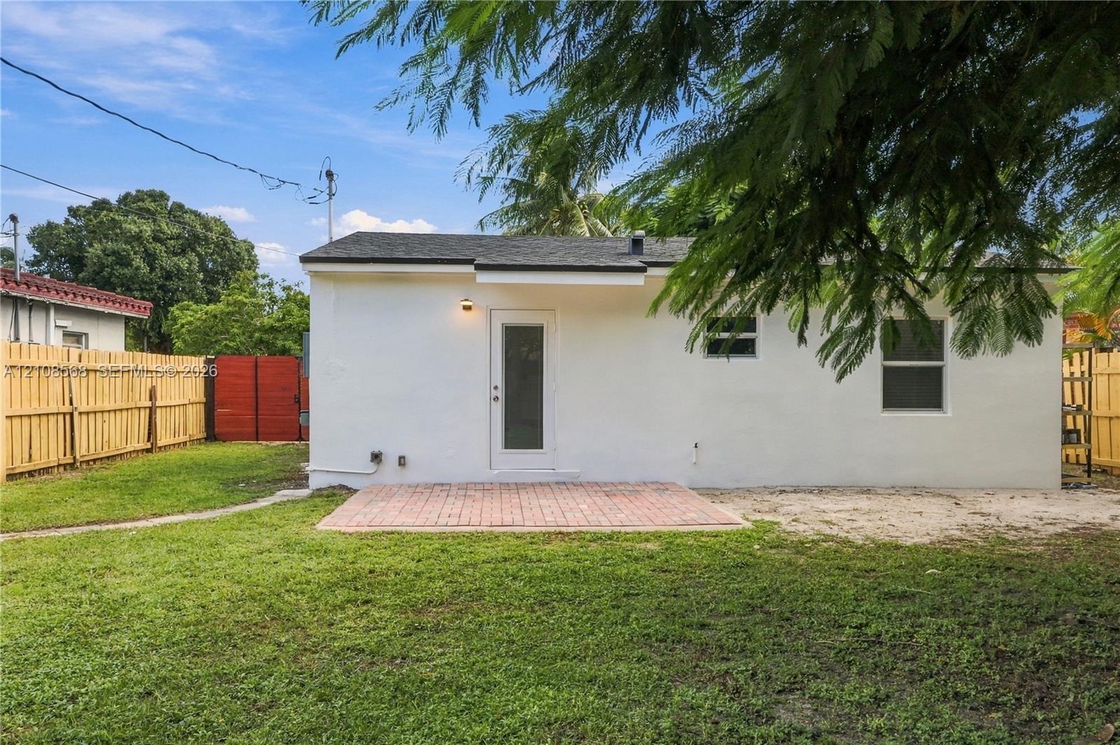 Exterior rear elevation featuring white stucco siding, a brick paver patio, full-height wooden privacy fencing, and a glass-panel exterior door