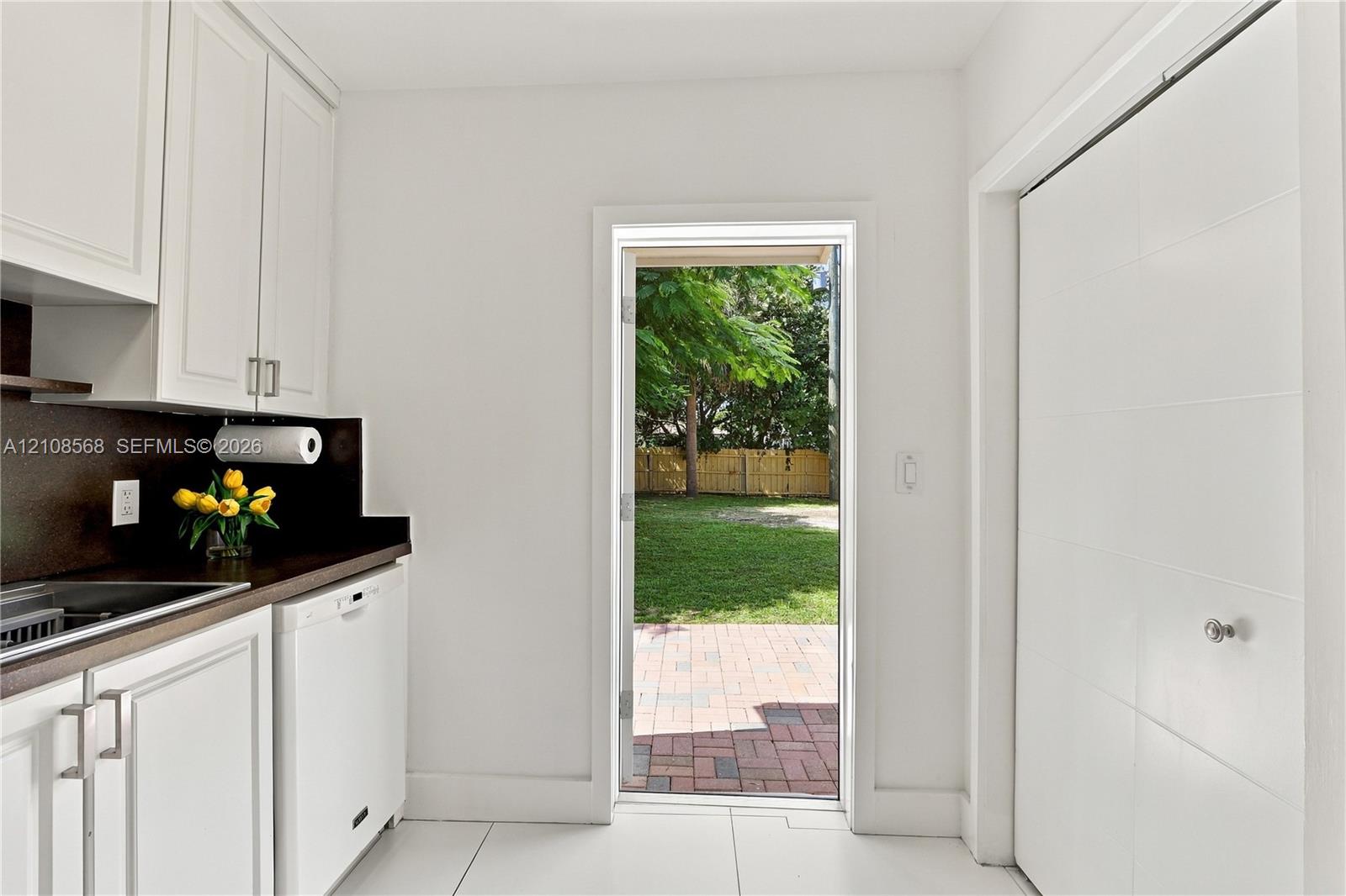 White cabinetry with modern hardware, contrasting dark countertops, built-in dishwasher, large-format tile flooring, and a glass-paneled door opening to a brick paver patio and lawn
