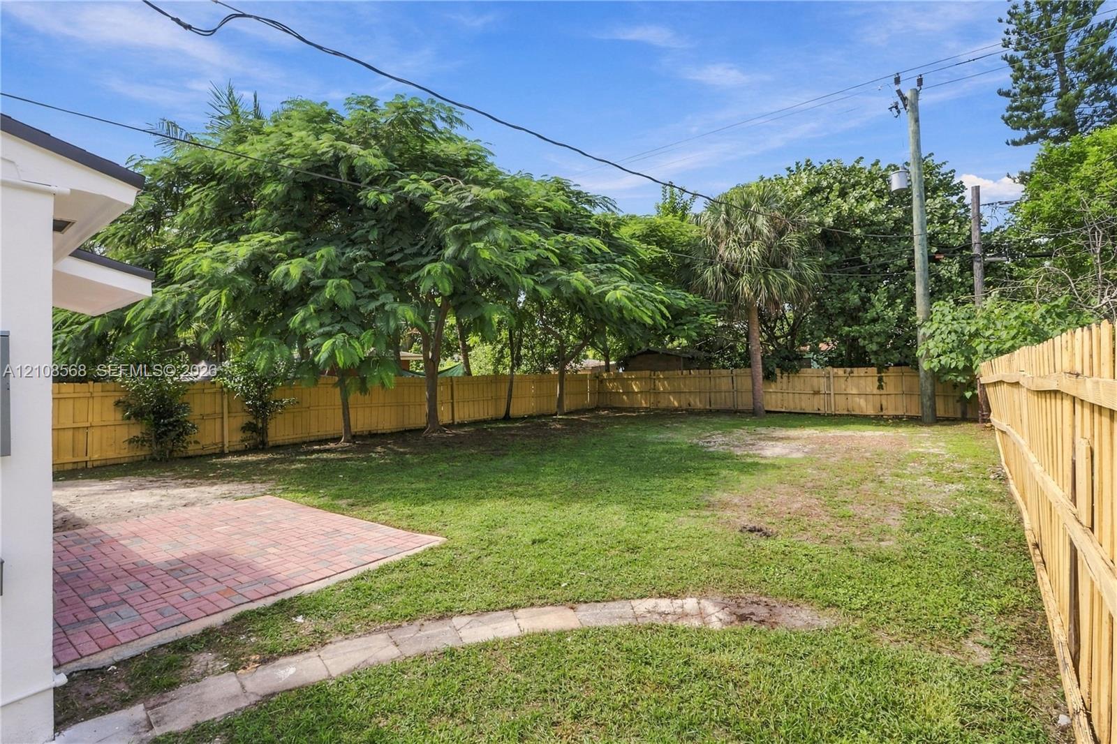 Backyard featuring a red brick patio, perimeter wood fencing, grass lawn, and mature trees